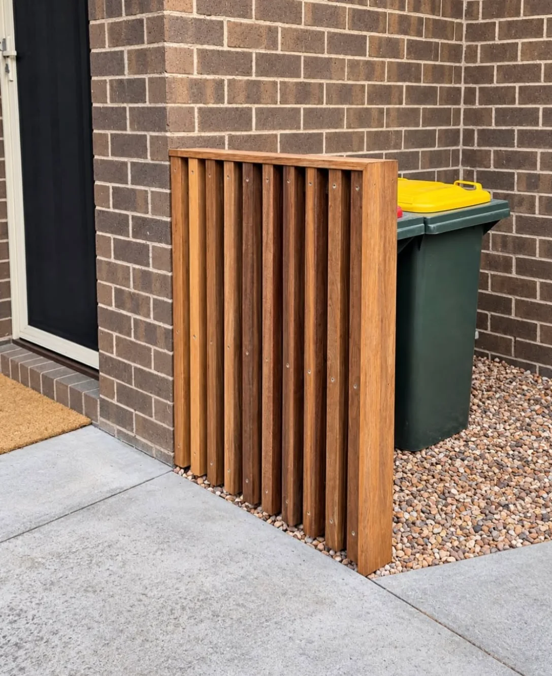 Custom hardwood bin screen with vertical timber battens beside a Port Melbourne front door, with a pebble base and grey paving